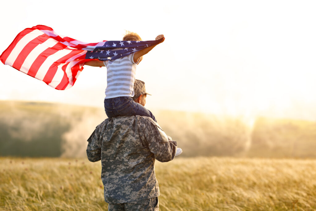 Child US flag on the shoulders of a miliary parent in a backyard or park demonstrates the importance of the VA Loan program for military veterans and their families.